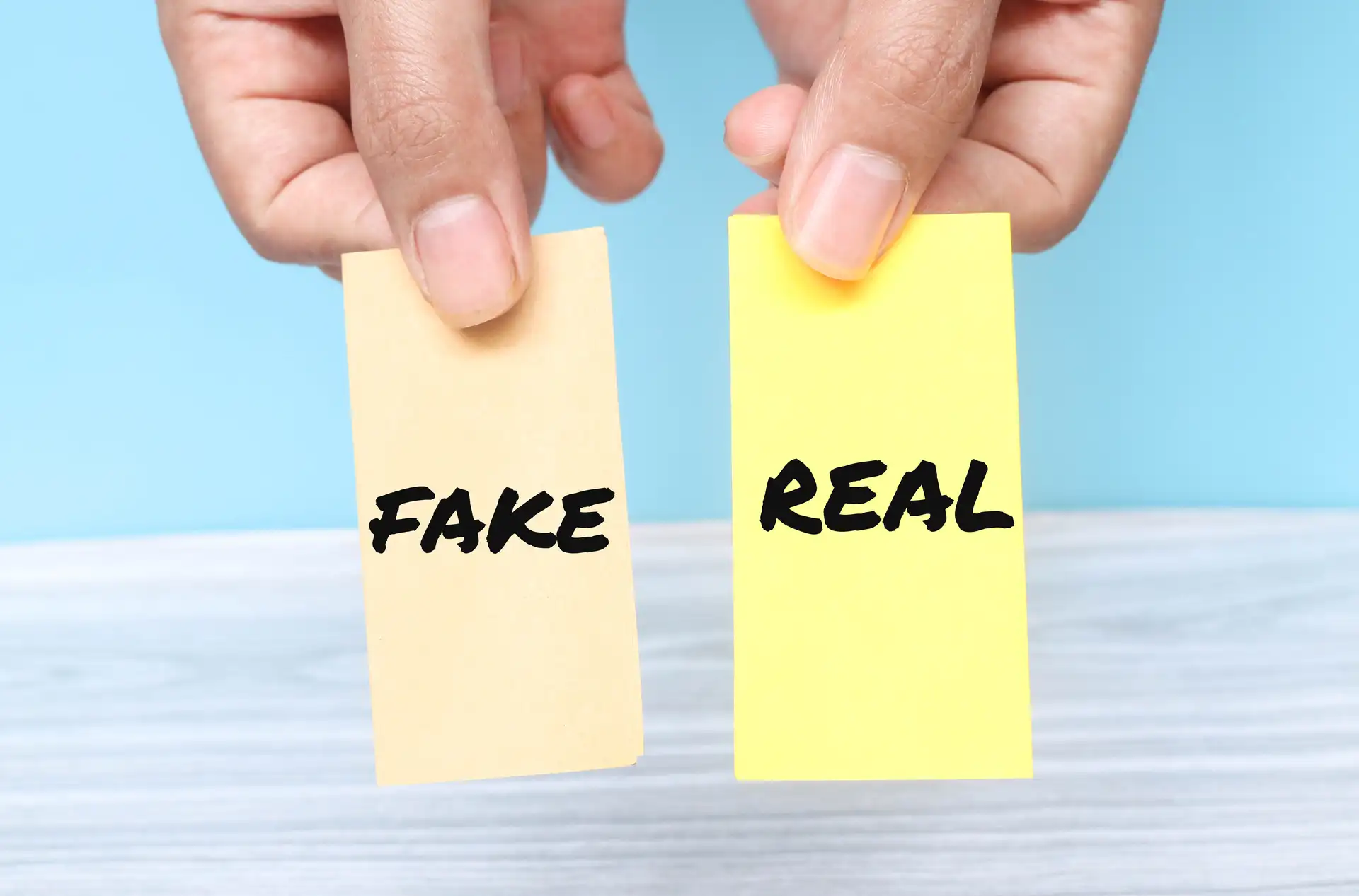 Two hands holding paper notes labelled FAKE and REAL against a soft blue background symbolising the choice between fake and genuine hookup profiles