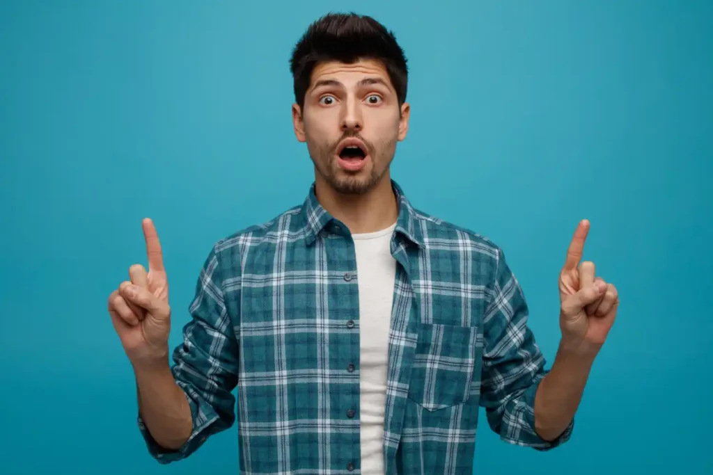 A young man in a checked shirt looking shocked and surprised with his mouth open and both index fingers pointing up, standing against a bright blue background, showing a reaction of surprise at unexpected bedroom advice